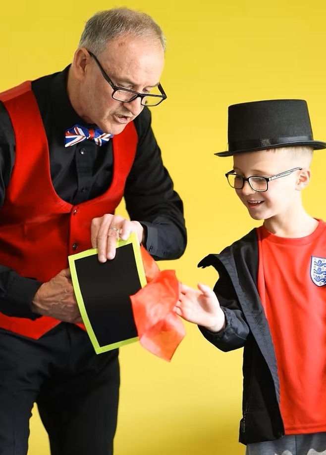 Michael Raw performing as Mr PunchandJudy, standing beside a traditional red‑and‑white Punch & Judy booth, smiling while entertaining children with puppets and balloon model